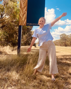 Sam stands in front of the Parkes shire sign with her arms in the air and smiling